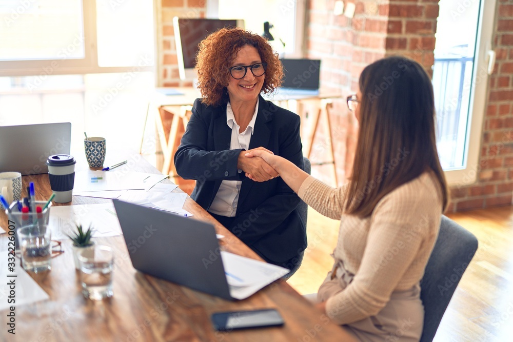 Fototapeta premium Two beautiful businesswomen smiling happy and confident. Sitting with smile on face working together using laptop shaking hands for agreement at the office