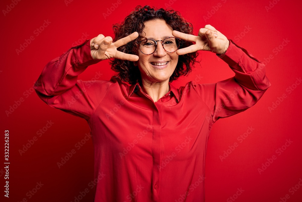 Middle age beautiful curly hair woman wearing casual shirt and glasses over red background Doing peace symbol with fingers over face, smiling cheerful showing victory