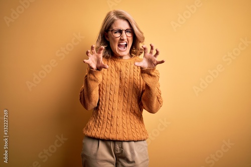 Middle age beautiful blonde woman wearing casual sweater and glasses over yellow background smiling funny doing claw gesture as cat, aggressive and sexy expression