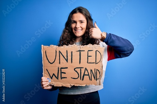 Beautiful activist woman holding banner with united stand message over blue background happy with big smile doing ok sign, thumb up with fingers, excellent sign © Krakenimages.com
