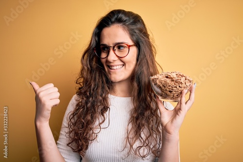 Young beautiful woman with curly hair holding bowl with healthy corn flakes cereals pointing and showing with thumb up to the side with happy face smiling