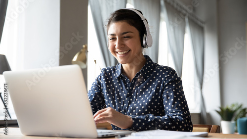 Smiling Indian girl wearing headphones using laptop, looking at screen, happy young female listening to favorite music while working online on project, excited student learning language photo