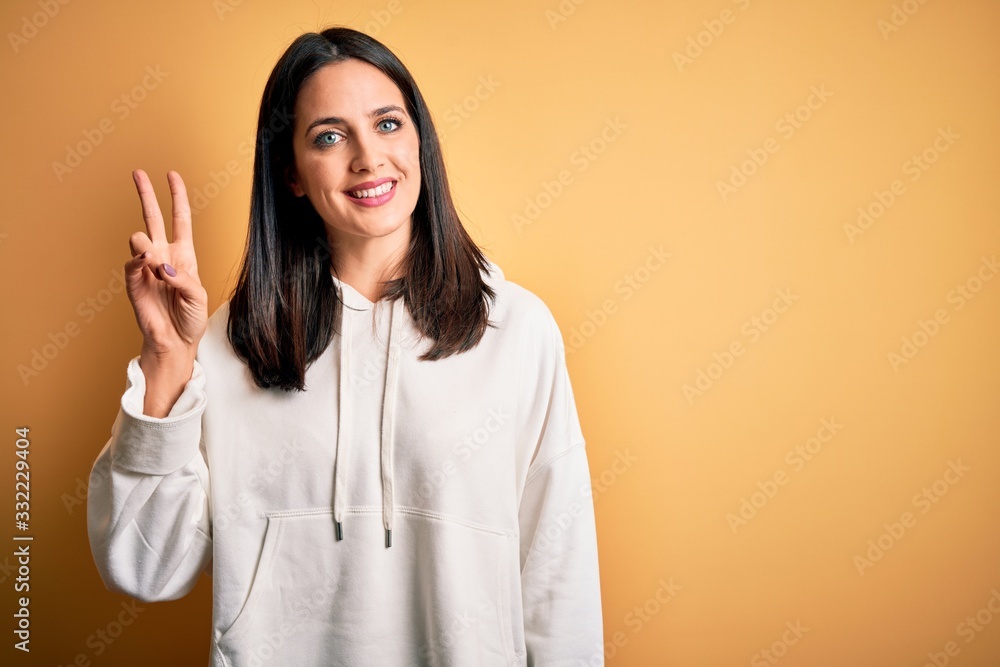 Young brunette sportswoman with blue eyes wearing training sweatshirt over yellow background smiling with happy face winking at the camera doing victory sign. Number two. Young brunette sportswoman with blue eyes wearing training sweatshirt over yellow background smiling with happy face winking at the camera doing victory sign. Number two.