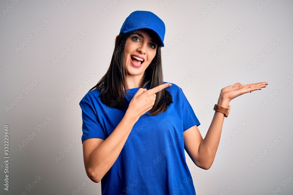 Young delivery woman with blue eyes wearing cap standing over blue background amazed and smiling to the camera while presenting with hand and pointing with finger. Young delivery woman with blue eyes wearing cap standing over blue background amazed and smiling to the camera while presenting with hand and pointing with finger.