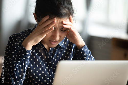 Unhappy Indian woman with closed eyes touching temples close up, feeling unwell, overworked female sitting at desk with laptop, upset exhausted girl suffering from strong headache or migraine