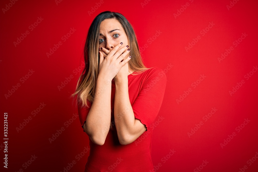 Young beautiful blonde woman with blue eyes wearing casual t-shirt over red background shocked covering mouth with hands for mistake. Secret concept. Young beautiful blonde woman with blue eyes wearing casual t-shirt over red background shocked covering mouth with hands for mistake. Secret concept.