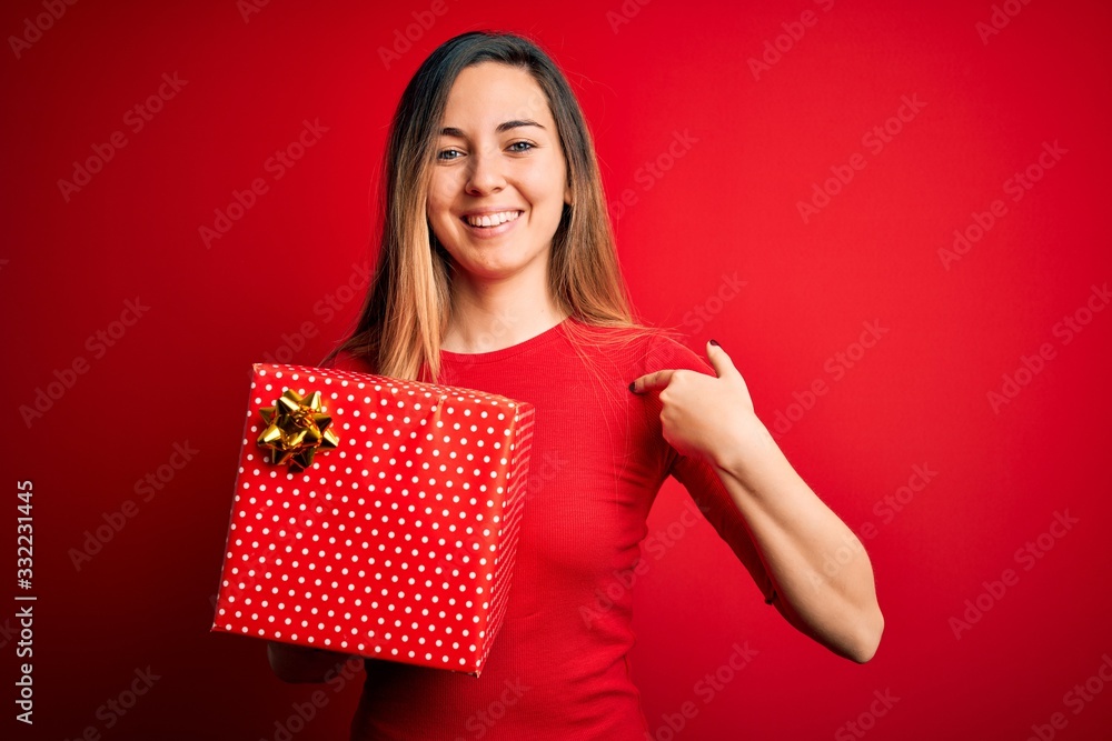 Young beautiful blonde woman with blue eyes holding birthday gift over red background with surprise face pointing finger to himself Young beautiful blonde woman with blue eyes holding birthday gift over red background with surprise face pointing finger to himself