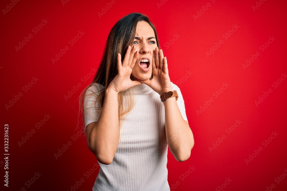 Fototapeta premium Beautiful blonde woman with blue eyes wearing casual white t-shirt over red background Shouting angry out loud with hands over mouth