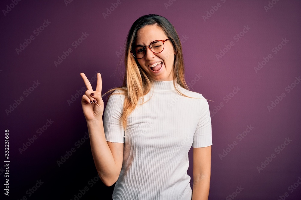 Young beautiful blonde woman with blue eyes wearing casual t-shirt over purple background smiling with happy face winking at the camera doing victory sign. Number two. Young beautiful blonde woman with blue eyes wearing casual t-shirt over purple background smiling with happy face winking at the camera doing victory sign. Number two.