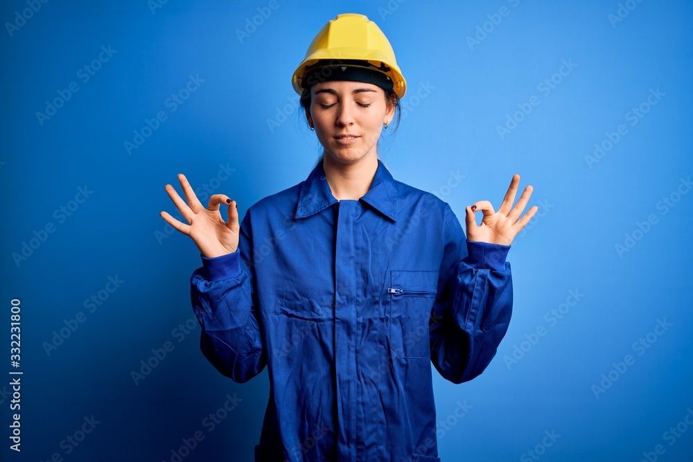 Young beautiful worker woman with blue eyes wearing security helmet and uniform relax and smiling with eyes closed doing meditation gesture with fingers. Yoga concept. Young beautiful worker woman with blue eyes wearing security helmet and uniform relax and smiling with eyes closed doing meditation gesture with fingers. Yoga concept.