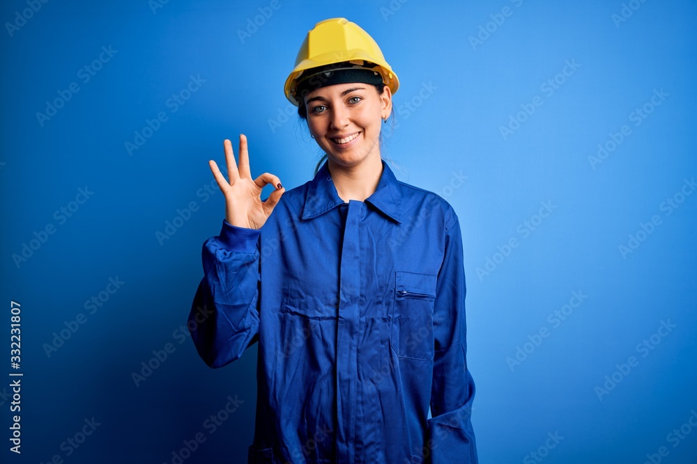 Young beautiful worker woman with blue eyes wearing security helmet and uniform smiling positive doing ok sign with hand and fingers. Successful expression.