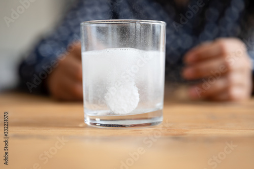 Close up sparkling water glass with dissolving effervescent aspirin pill standing on wooden table, sick unhealthy woman taking emergency medicine, painkiller to relieve headache or fever