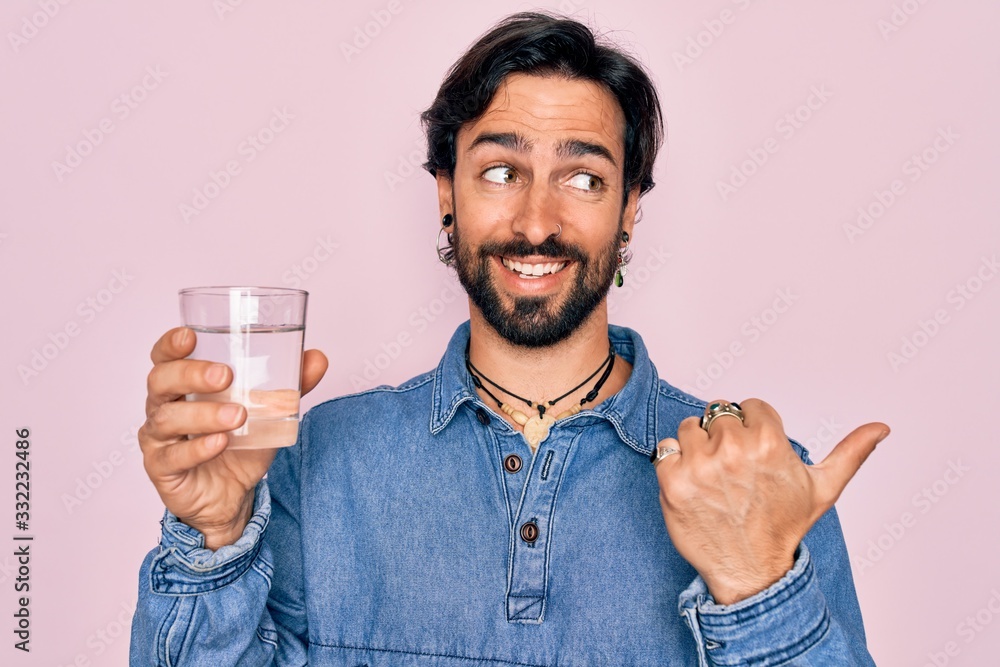 Young handsome hispanic bohemian hipster man with bear drinking a fresh glass of water pointing and showing with thumb up to the side with happy face smiling Young handsome hispanic bohemian hipster man with bear drinking a fresh glass of water pointing and showing with thumb up to the side with happy face smiling