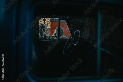 Two persons in NBC protective suits and gas masks, view through cabin of old truck.