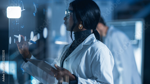 In the Research Laboratory Smart and Beautiful African American Female Scientist Wearing White Coat and Protective Glasses Writes Formula on Glass Whiteboard, References Her Tablet Computer