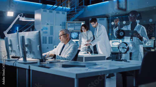 Diverse International Team of Industrial Scientists and Engineers Wearing White Coats Working on Industrial Machinery Design in Research Laboratory. Professionals Using Computers and Talking