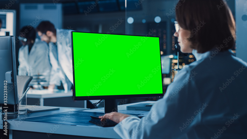 Over the Shoulder Shot: Female IT Scientist Uses Green Mock-up Screen Computer. In the Background Technology Research And Development Laboratory with Scientists, Engineers Working on High Tech Design Over the Shoulder Shot: Female IT Scientist Uses Green Mock-up Screen Computer. In the Background Technology Research And Development Laboratory with Scientists, Engineers Working on High Tech Design