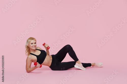 Fitness girl with perfect slim and fit body training muscles with dumbbells on pink background. Sporty woman in sportswear