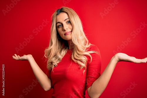 Young beautiful blonde woman wearing casual t-shirt standing over isolated red background clueless and confused expression with arms and hands raised. Doubt concept. © Krakenimages.com