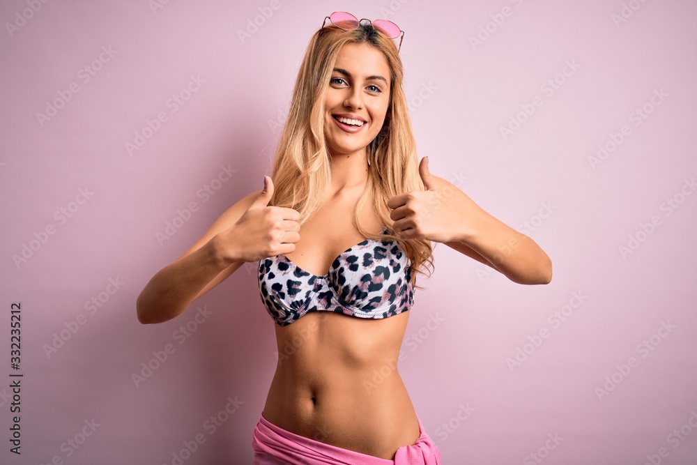 Obraz premium Young beautiful blonde woman on vacation wearing bikini over isolated pink background success sign doing positive gesture with hand, thumbs up smiling and happy. Cheerful expression and winner