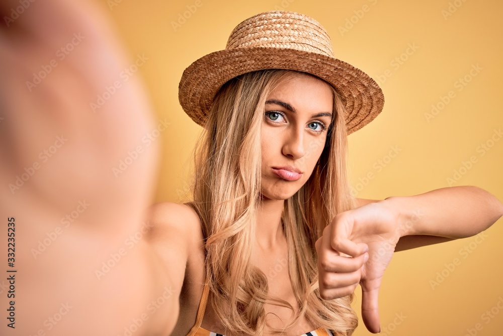 Young beautiful blonde woman on vacation wearing bikini and hat making selfie by camera with angry face, negative sign showing dislike with thumbs down, rejection concept Young beautiful blonde woman on vacation wearing bikini and hat making selfie by camera with angry face, negative sign showing dislike with thumbs down, rejection concept