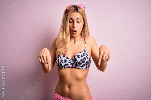 Young beautiful blonde woman on vacation wearing bikini over isolated pink background Pointing down with fingers showing advertisement, surprised face and open mouth © Krakenimages.com