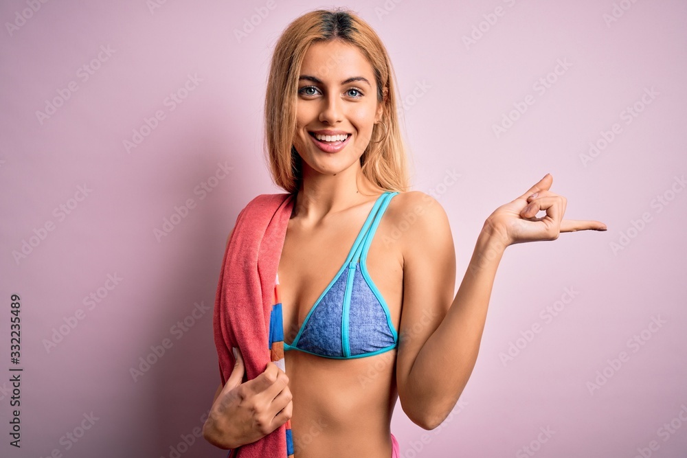 Beautiful blonde woman on vacation wearing bikini holding beach towel over pink background very happy pointing with hand and finger to the side Beautiful blonde woman on vacation wearing bikini holding beach towel over pink background very happy pointing with hand and finger to the side