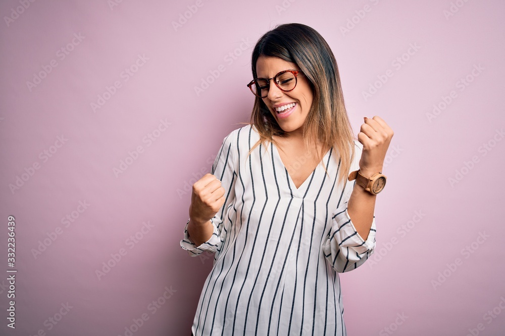 Young beautiful woman wearing casual striped t-shirt and glasses over pink background celebrating surprised and amazed for success with arms raised and eyes closed. Winner concept. Young beautiful woman wearing casual striped t-shirt and glasses over pink background celebrating surprised and amazed for success with arms raised and eyes closed. Winner concept.