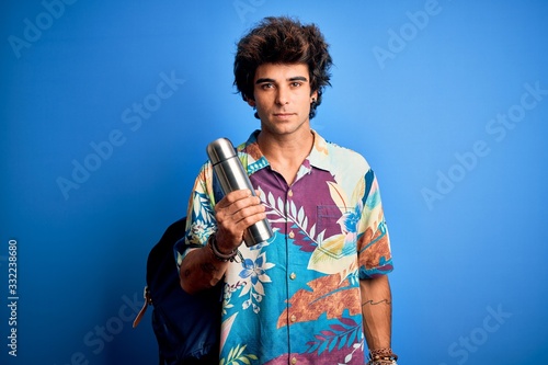 Young tourist man wearing backpack holding thermo over isolated blue background with a confident expression on smart face thinking serious