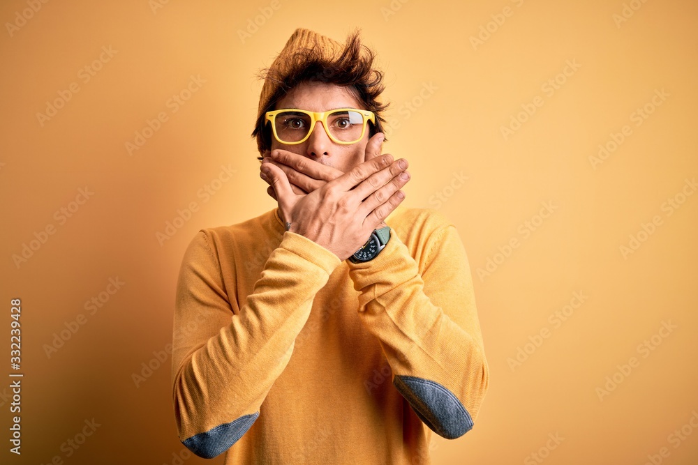 Young handsome man wearing casual t-shirt and glasses over isolated yellow background shocked covering mouth with hands for mistake. Secret concept.