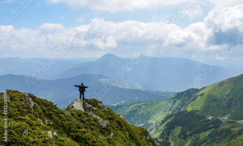  Hiker with his arms spread to the side standing on top of mountain ridge overlooking peaked mountains and forest valley against cloudy sky and enjoying the freedom and view