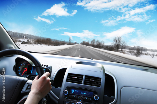 Car dashboard with driver's hand on the steering wheel against a winter road in motion and sky with clouds