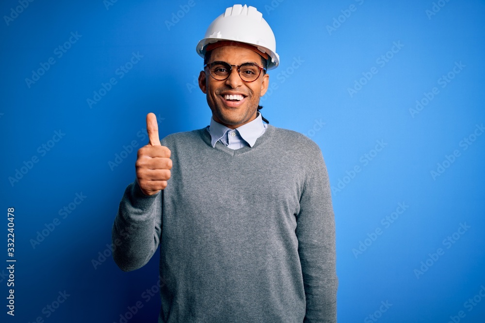 Young handsome african american engineer man with dreadlocks wearing safety helmet doing happy thumbs up gesture with hand. Approving expression looking at the camera showing success.