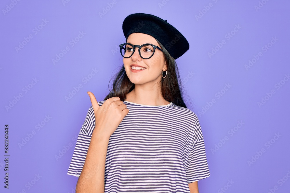 Young beautiful brunette woman wearing glasses and french beret over purple background smiling with happy face looking and pointing to the side with thumb up. Young beautiful brunette woman wearing glasses and french beret over purple background smiling with happy face looking and pointing to the side with thumb up.