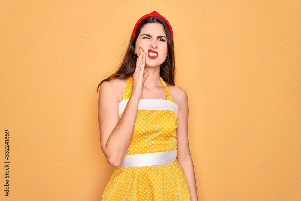 Young beautiful pin up woman wearing 50s fashion vintage dress over yellow background touching mouth with hand with painful expression because of toothache or dental illness on teeth. Dentist Young beautiful pin up woman wearing 50s fashion vintage dress over yellow background touching mouth with hand with painful expression because of toothache or dental illness on teeth. Dentist