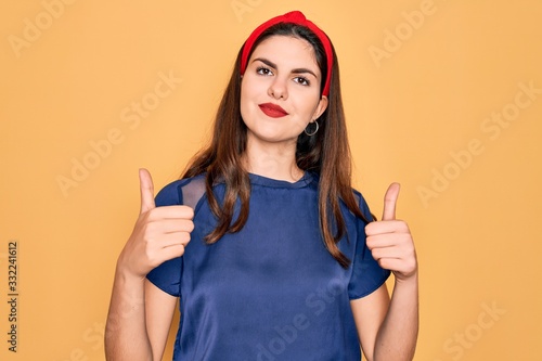 Young beautiful brunette woman wearing red lips over yellow background success sign doing positive gesture with hand, thumbs up smiling and happy. Cheerful expression and winner gesture.
