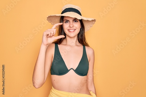 Young beautiful girl wearing swimwear bikini and summer sun hat over yellow background smiling and confident gesturing with hand doing small size sign with fingers looking and the camera. Measure