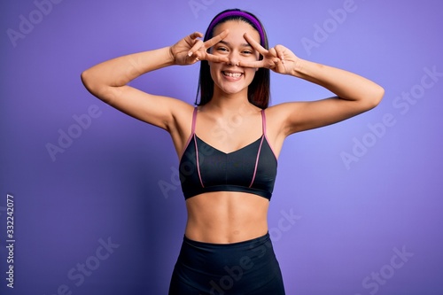 Young beautiful sporty girl doing sport wearing sportswear over isolated purple background Doing peace symbol with fingers over face, smiling cheerful showing victory © Krakenimages.com