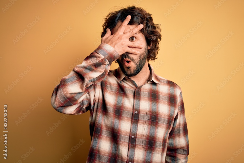 Young handsome man with beard wearing casual shirt standing over yellow background peeking in shock covering face and eyes with hand, looking through fingers with embarrassed expression. Young handsome man with beard wearing casual shirt standing over yellow background peeking in shock covering face and eyes with hand, looking through fingers with embarrassed expression.