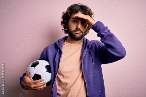 Handsome player man with beard playing soccer holding football ball over pink background stressed with hand on head, shocked with shame and surprise face, angry and frustrated. Fear and upset © Krakenimages.com