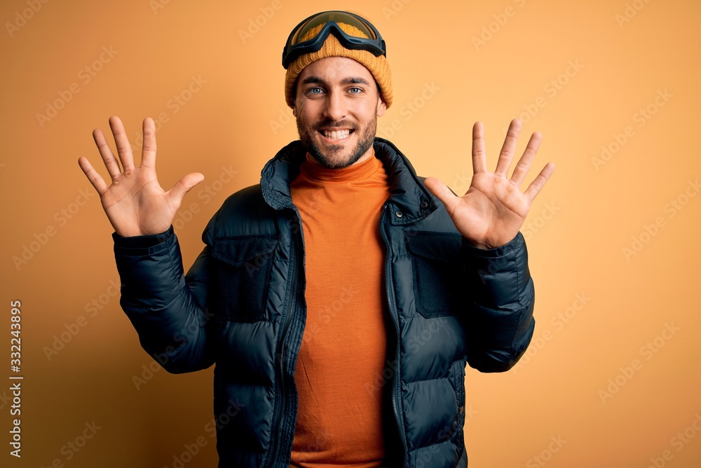 Young handsome skier man with beard wearing snow sportswear and ski goggles showing and pointing up with fingers number ten while smiling confident and happy. Young handsome skier man with beard wearing snow sportswear and ski goggles showing and pointing up with fingers number ten while smiling confident and happy.