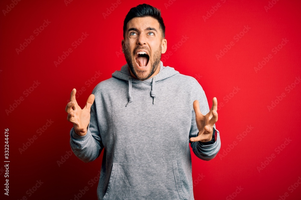 Young handsome sportsman wearing sweatshirt standing over isolated red background crazy and mad shouting and yelling with aggressive expression and arms raised. Frustration concept. Young handsome sportsman wearing sweatshirt standing over isolated red background crazy and mad shouting and yelling with aggressive expression and arms raised. Frustration concept.