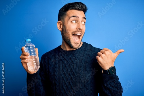 Young handsome man drinking bottle of water to refeshment over blue background pointing and showing with thumb up to the side with happy face smiling photo