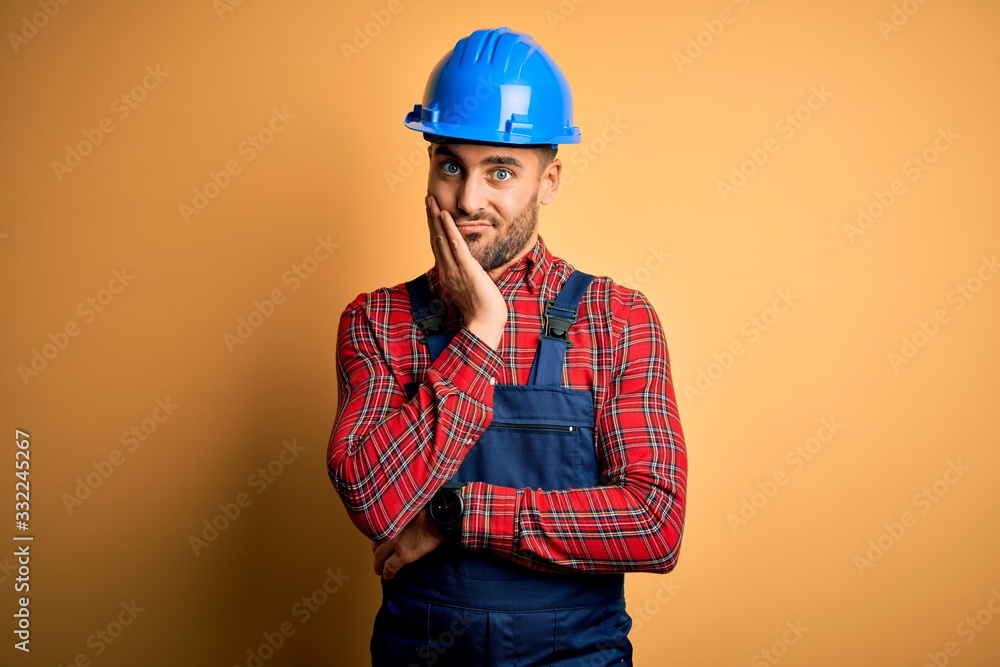 Young builder man wearing construction uniform and safety helmet over yellow background thinking looking tired and bored with depression problems with crossed arms.