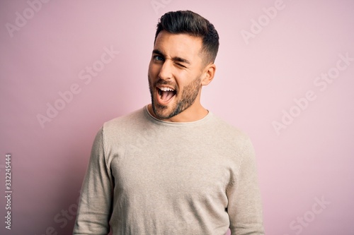 Young handsome man wearing casual sweater standing over isolated pink background winking looking at the camera with sexy expression, cheerful and happy face.