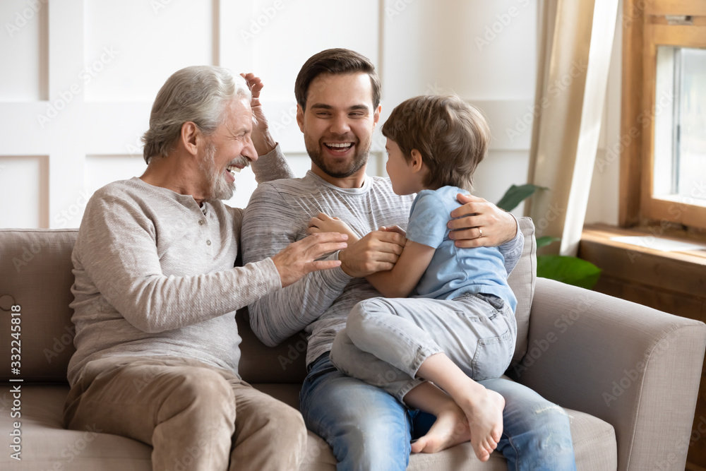 Happy three generations of men sit relax on couch in living room having fun together, overjoyed small preschooler boy play with young dad and elderly grandfather, enjoy family weekend at home