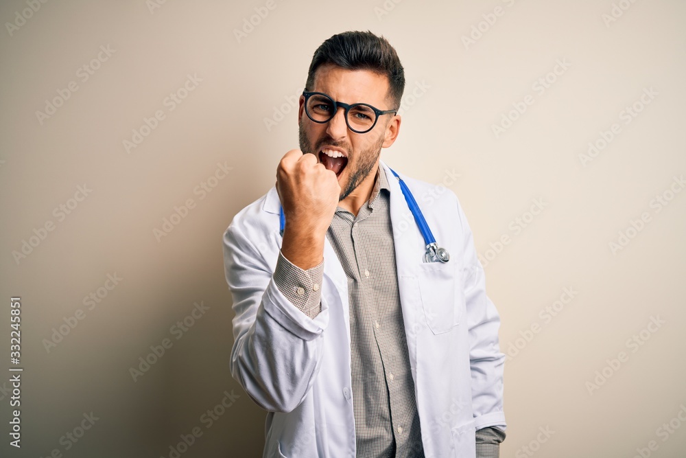 Young doctor man wearing glasses, medical white robe and stethoscope over isolated background angry and mad raising fist frustrated and furious while shouting with anger. Rage and aggressive concept. Young doctor man wearing glasses, medical white robe and stethoscope over isolated background angry and mad raising fist frustrated and furious while shouting with anger. Rage and aggressive concept.