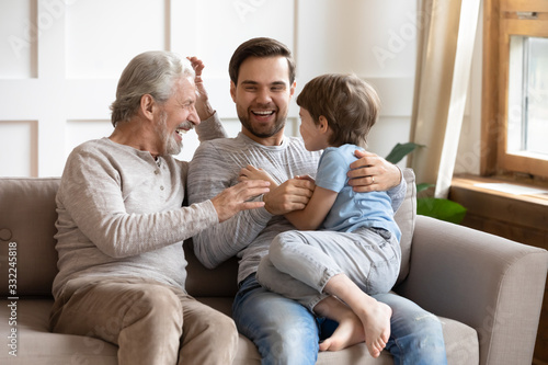 Happy three generations of men sit relax on couch in living room having fun together, overjoyed small preschooler boy play with young dad and elderly grandfather, enjoy family weekend at home