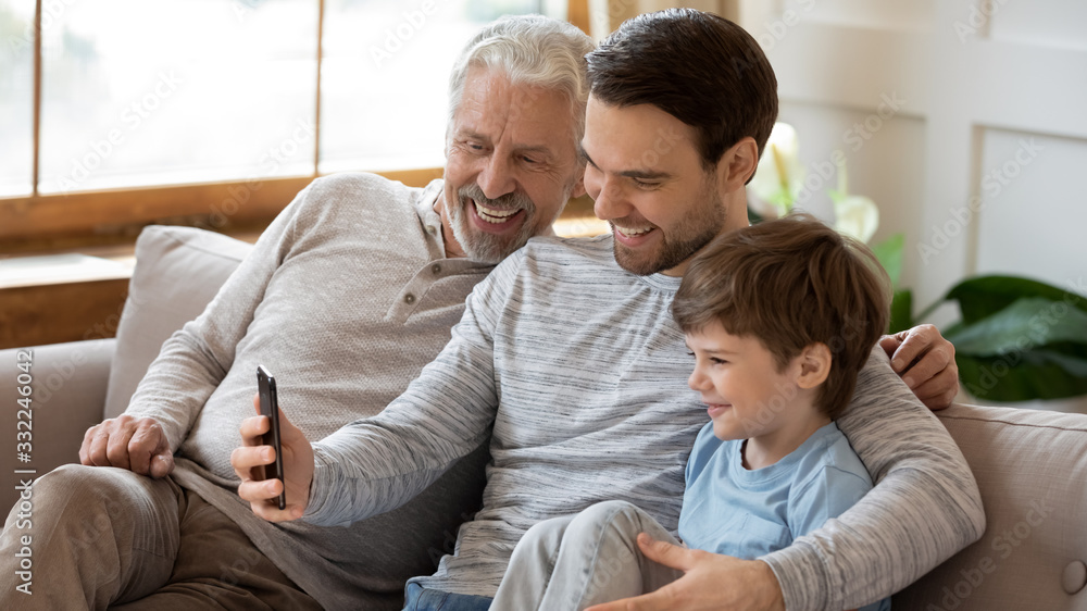 Smiling three generations of men sit on couch in living room make self-portrait picture on smartphone, happy little boy with young dad and mature grandfather take selfie on cellphone gadget together