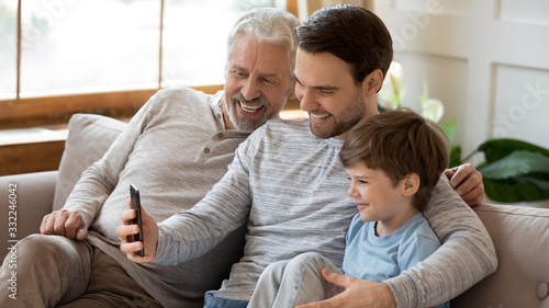 Smiling three generations of men sit on couch in living room make self-portrait picture on smartphone, happy little boy with young dad and mature grandfather take selfie on cellphone gadget together